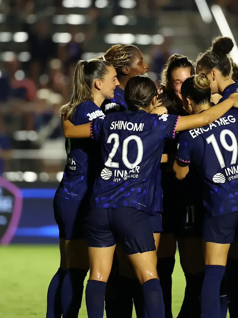 Women's football team in a tight celebratory huddle on the pitch at night